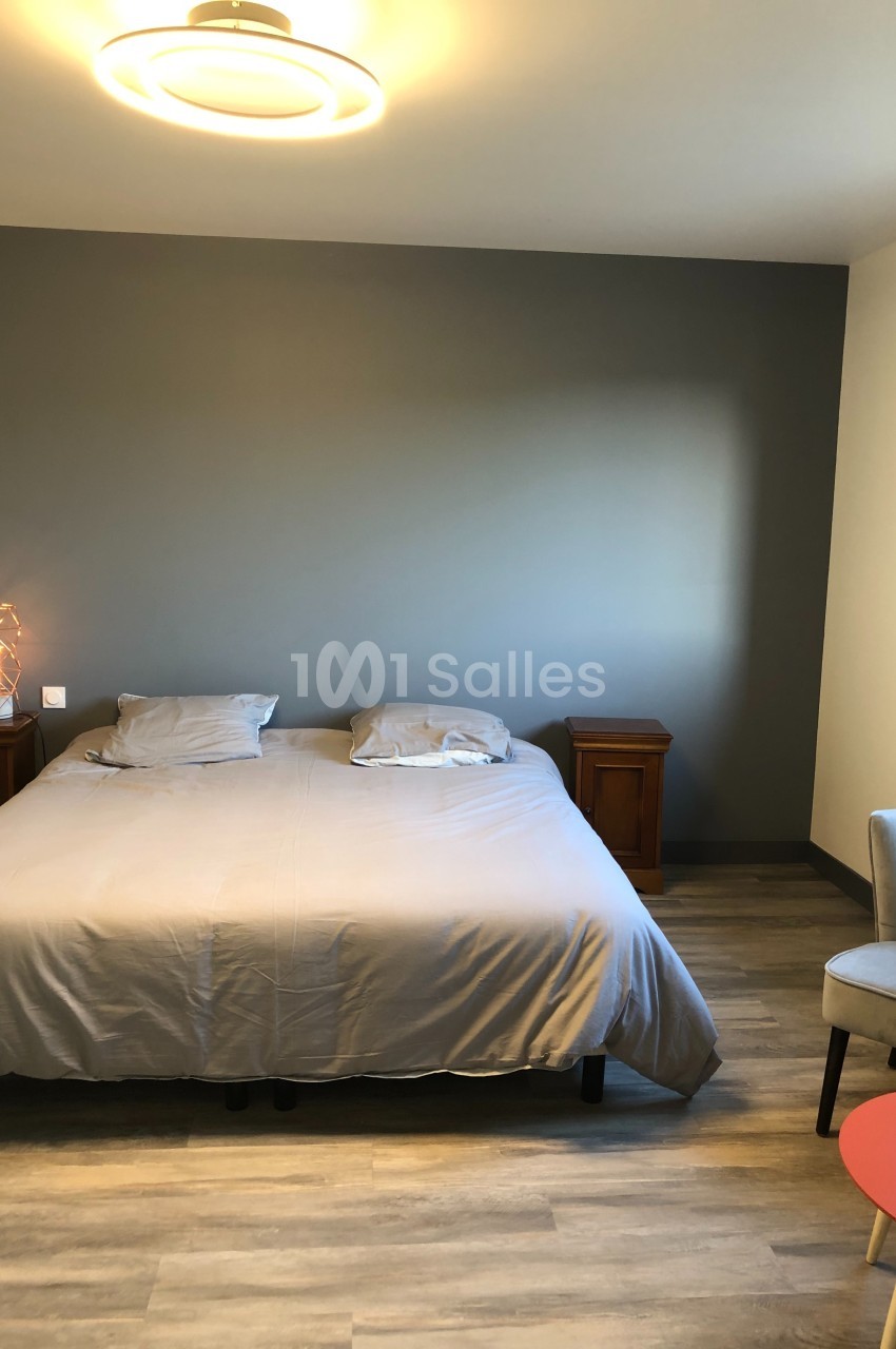 Chambre avec un lit double, draps gris, deux tables de chevet en bois, mur gris et sol en parquet clair.