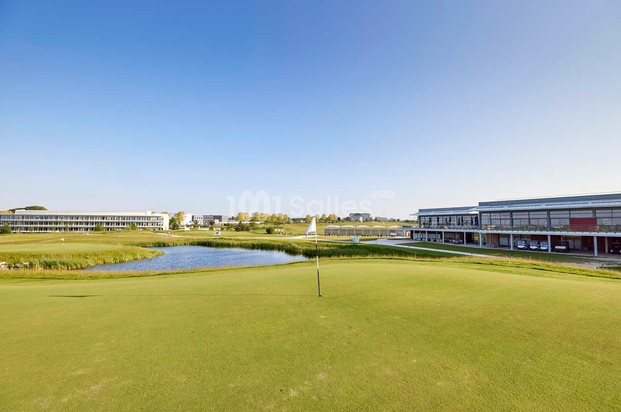 Vue d'un terrain de golf avec un drapeau, un étang et des bâtiments modernes en arrière-plan sous un ciel dégagé.