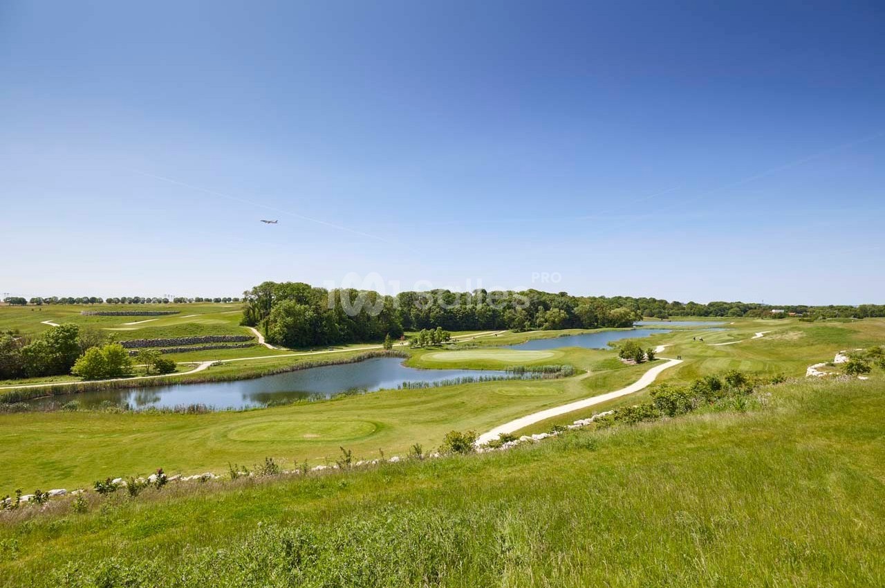 Paysage de golf avec étangs, pelouses verdoyantes, sentiers sinueux et arbres sous un ciel bleu dégagé.
