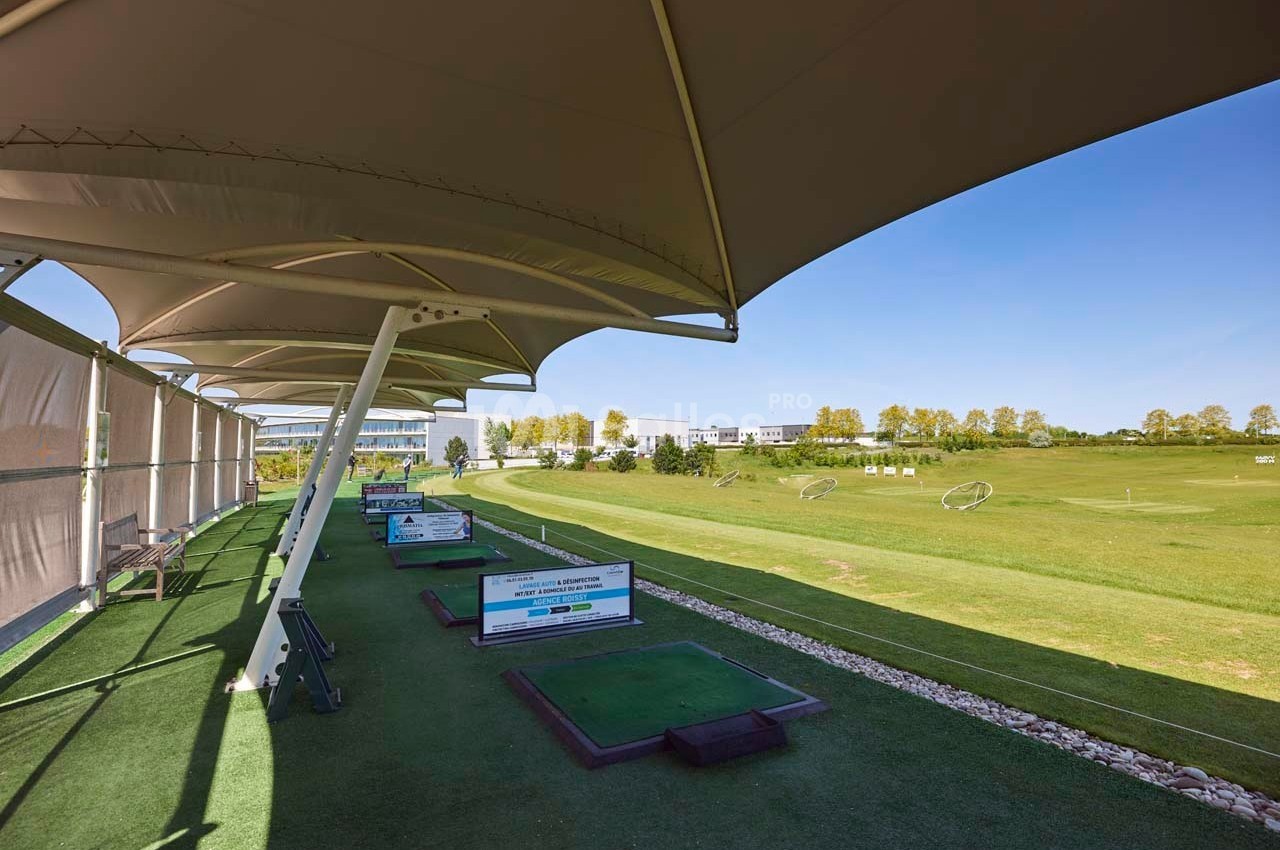 Poste de practice de golf couvert avec tapis d'entraînement, vue sur un terrain dégagé et ciel bleu.
