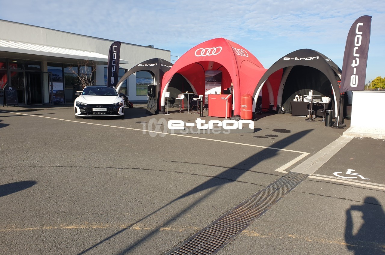 Stand extérieur avec tentes rouges et noires, logo ’e-tron’, voiture exposée devant un bâtiment moderne.
