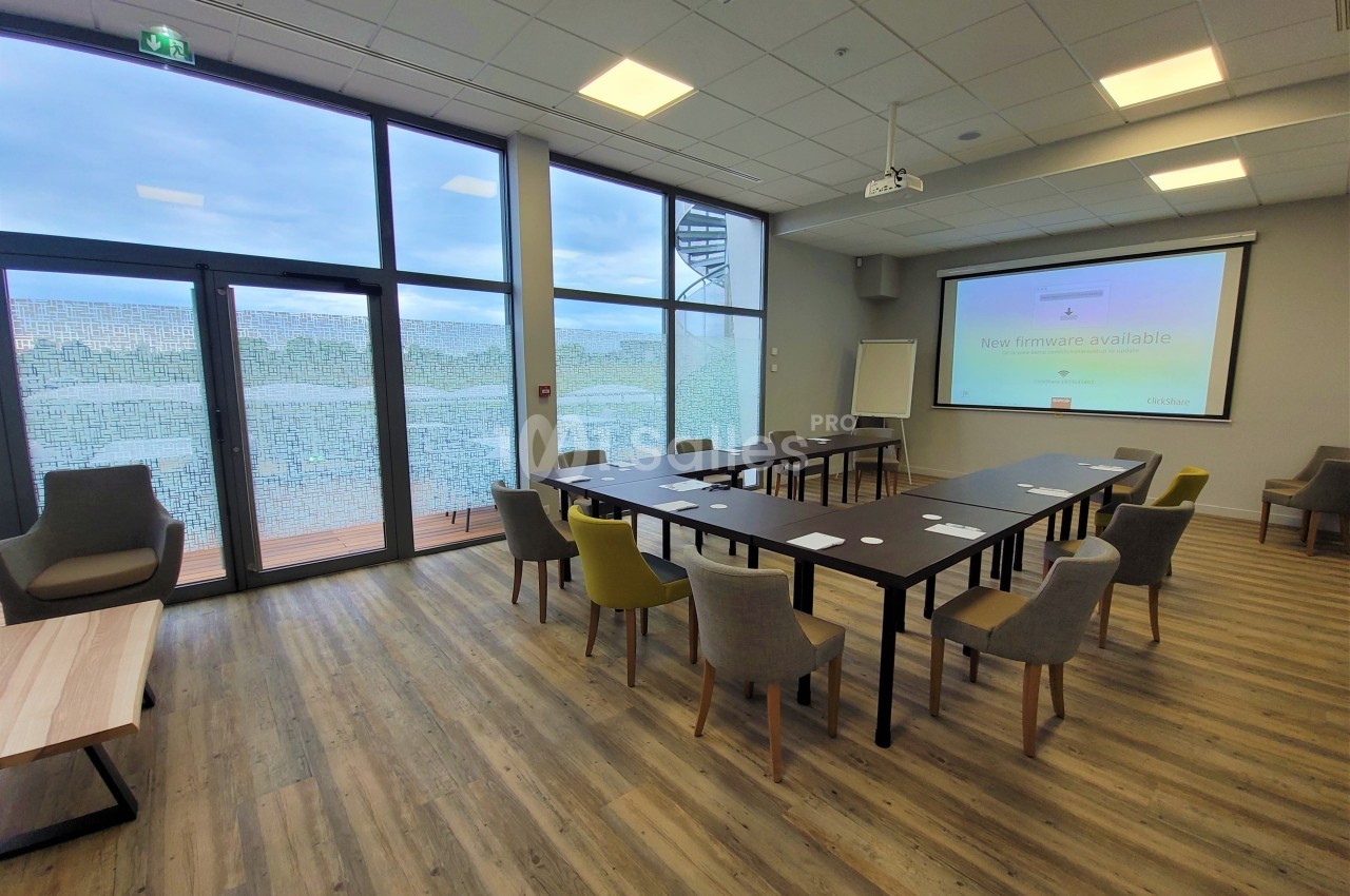Salle de réunion lumineuse avec grandes baies vitrées, tables disposées en U et chaises de différentes couleurs.