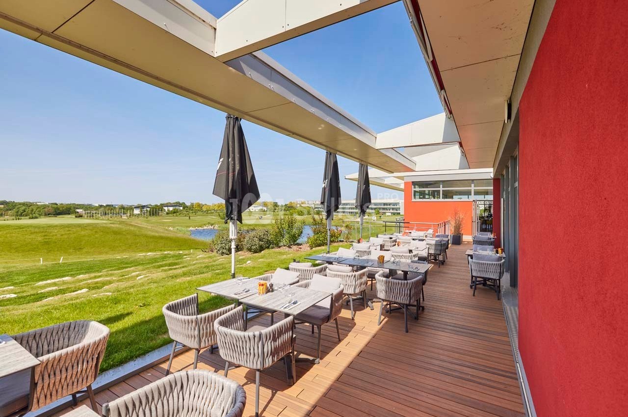 Terrasse en bois avec tables et chaises, surplombant un terrain de golf verdoyant par une journée ensoleillée.