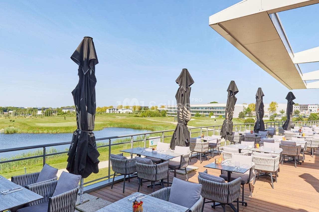 Terrasse d'un restaurant avec tables et parasols, offrant une vue sur un lac et un paysage verdoyant par temps ensoleillé.