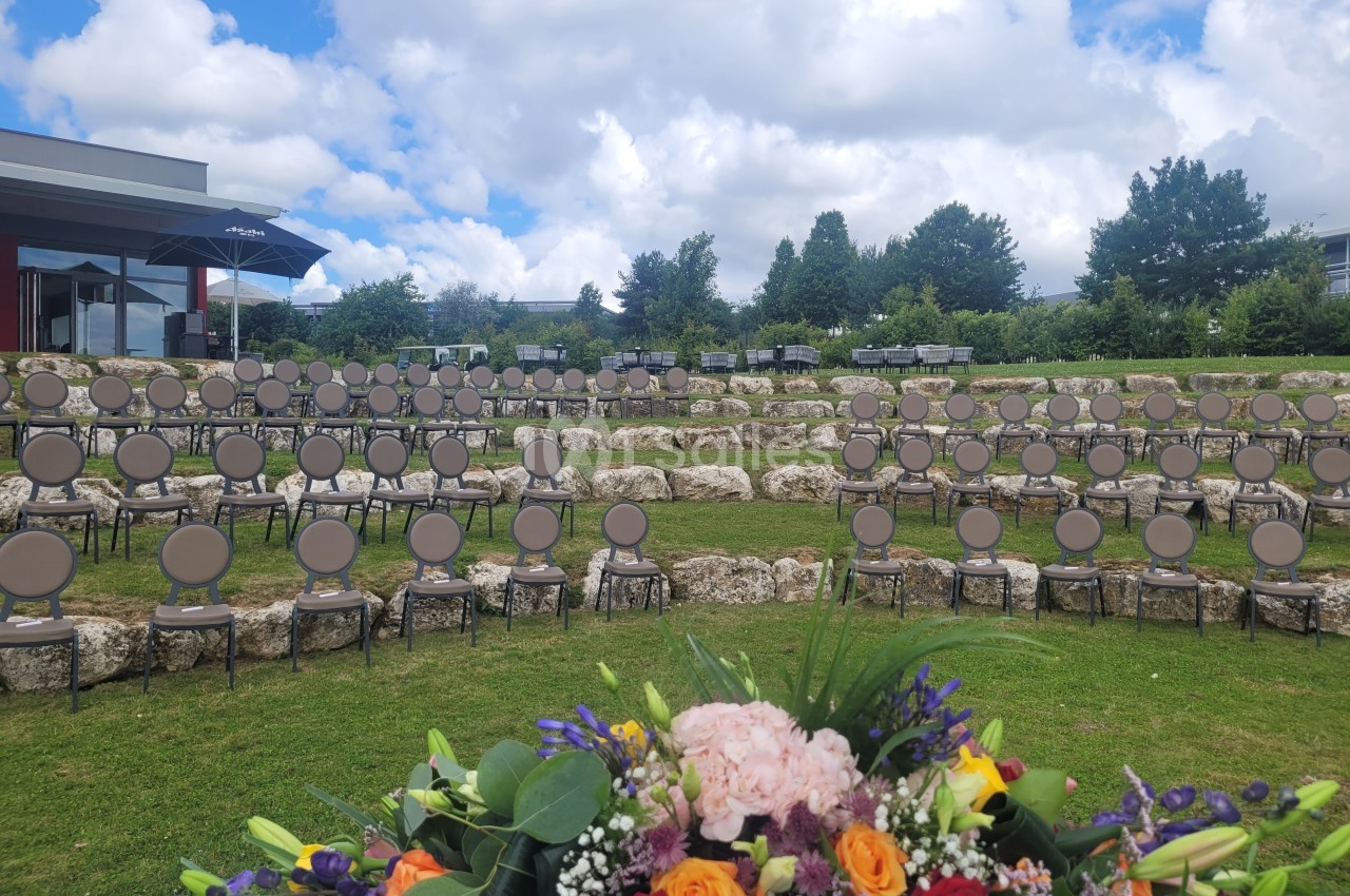 Chaises disposées en gradins sur une pelouse, avec un bouquet de fleurs au premier plan et un ciel nuageux en arrière-plan.