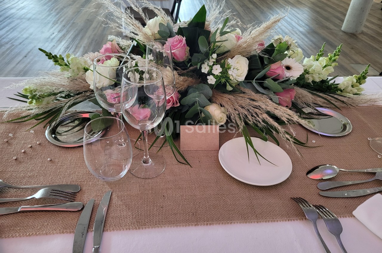 Centre de table avec fleurs roses et blanches, verres à vin, assiettes et couverts disposés sur une nappe beige.