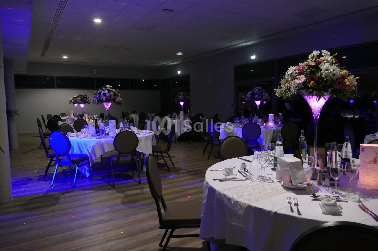 Salle de réception élégamment décorée avec des tables rondes, nappes blanches et centres de table lumineux.