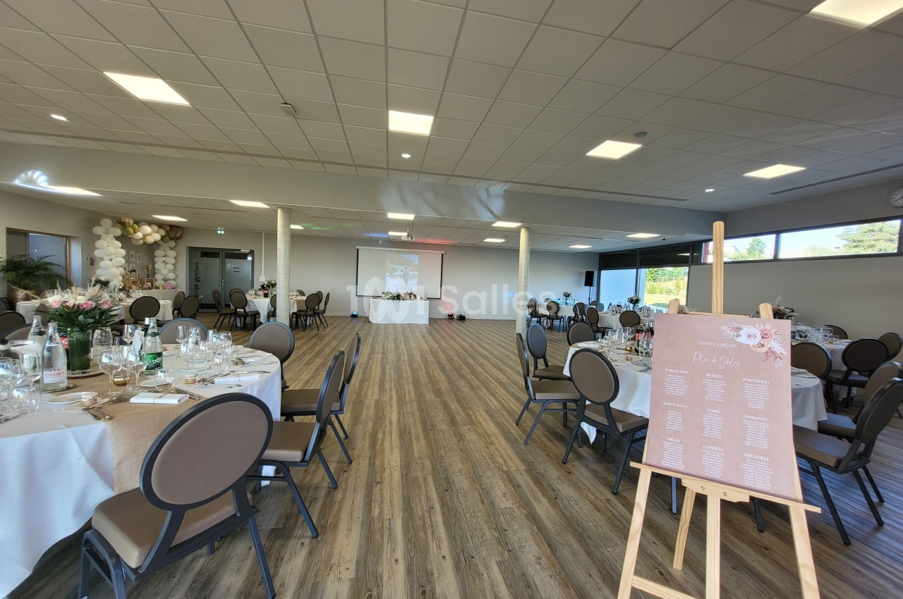 Salle de réception lumineuse avec tables dressées pour un événement et un plan de table sur un chevalet au premier plan.