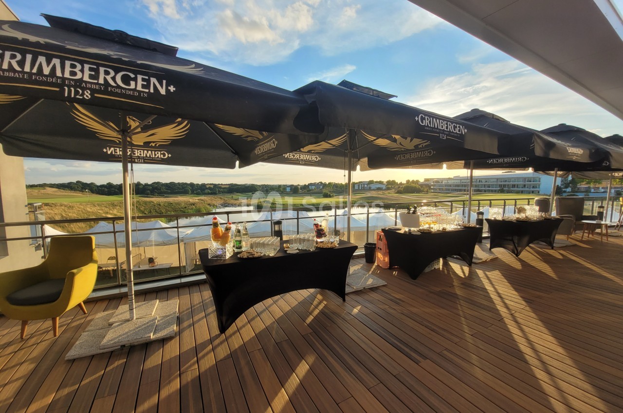 Terrasse en bois avec tables dressées sous des parasols noirs, offrant une vue sur un paysage naturel au coucher du soleil.