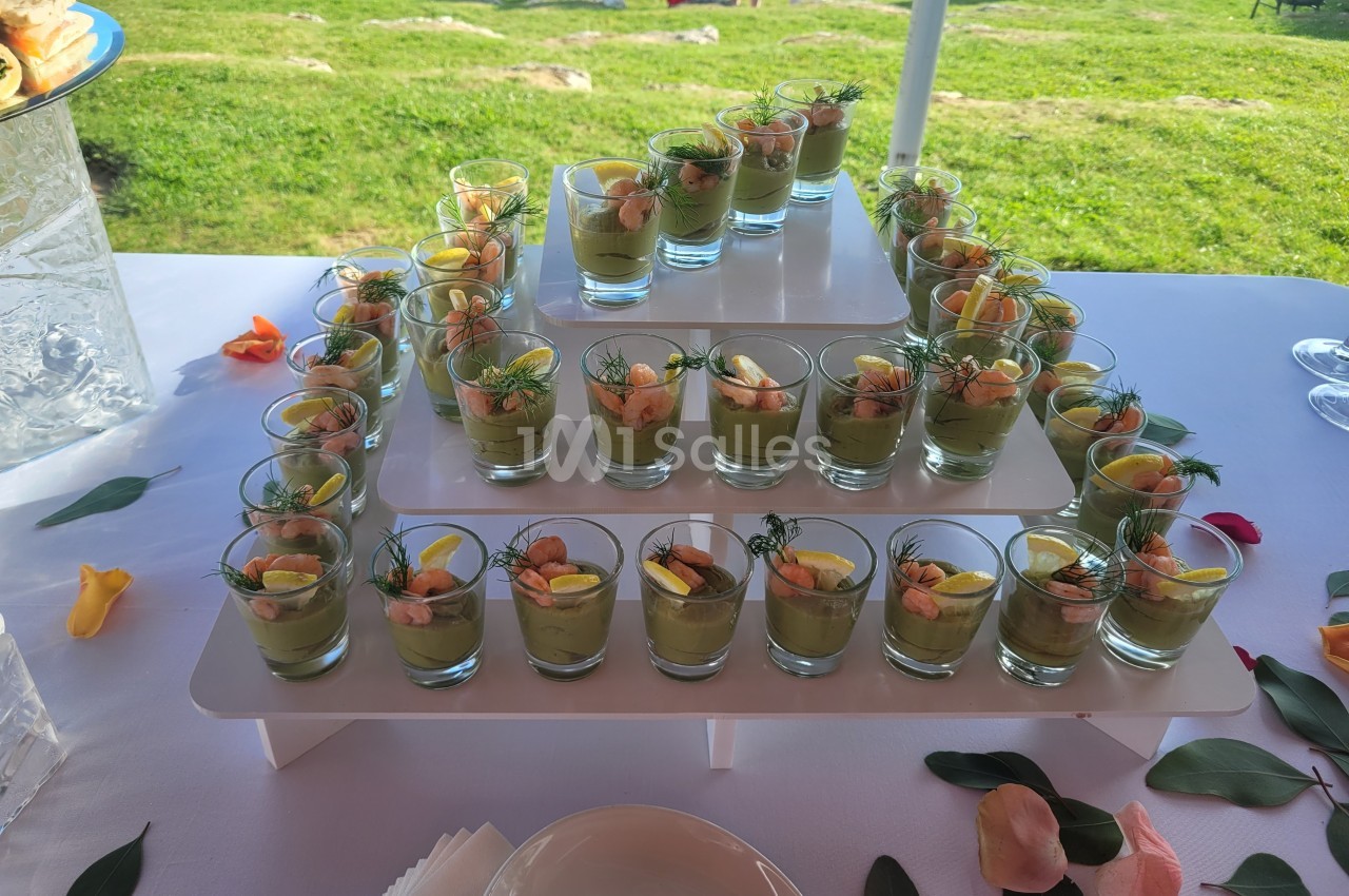 Présentoir de verrines garnies de mousse verte, crevettes et herbes fraîches, disposé sur une table en extérieur.