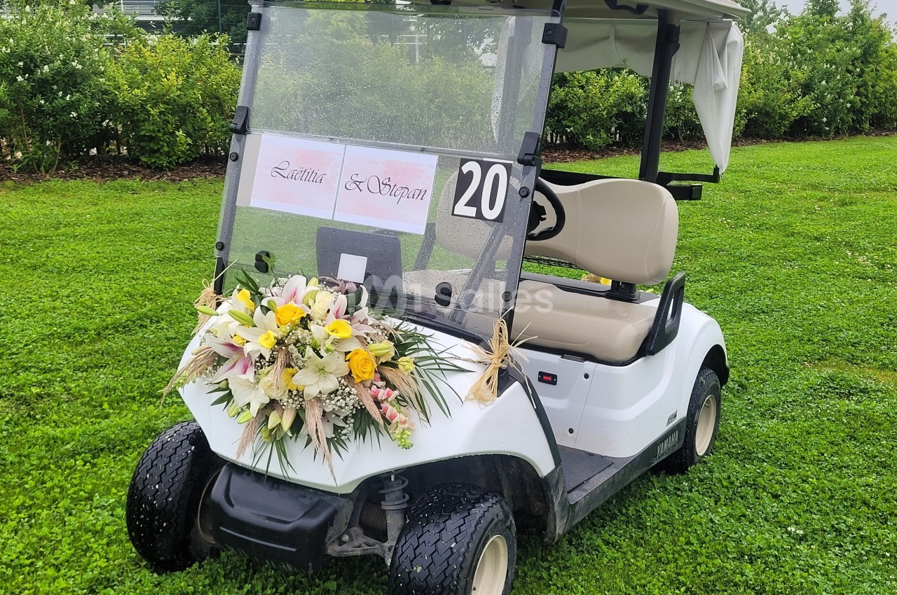 Voiturette de golf blanche décorée de fleurs et d'un panneau avec des prénoms, stationnée sur une pelouse verte.