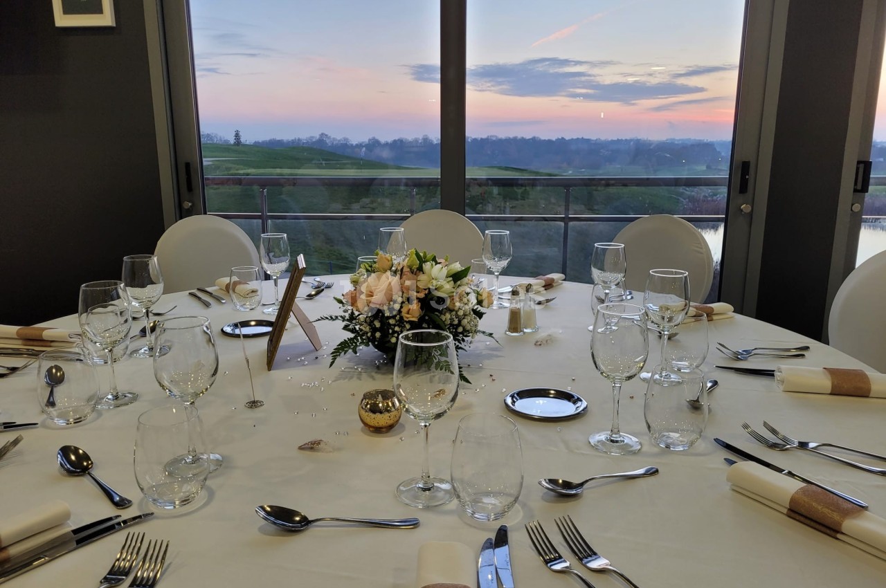 Table élégamment dressée avec des couverts, verres et centre de table floral, devant une baie vitrée au coucher du soleil.