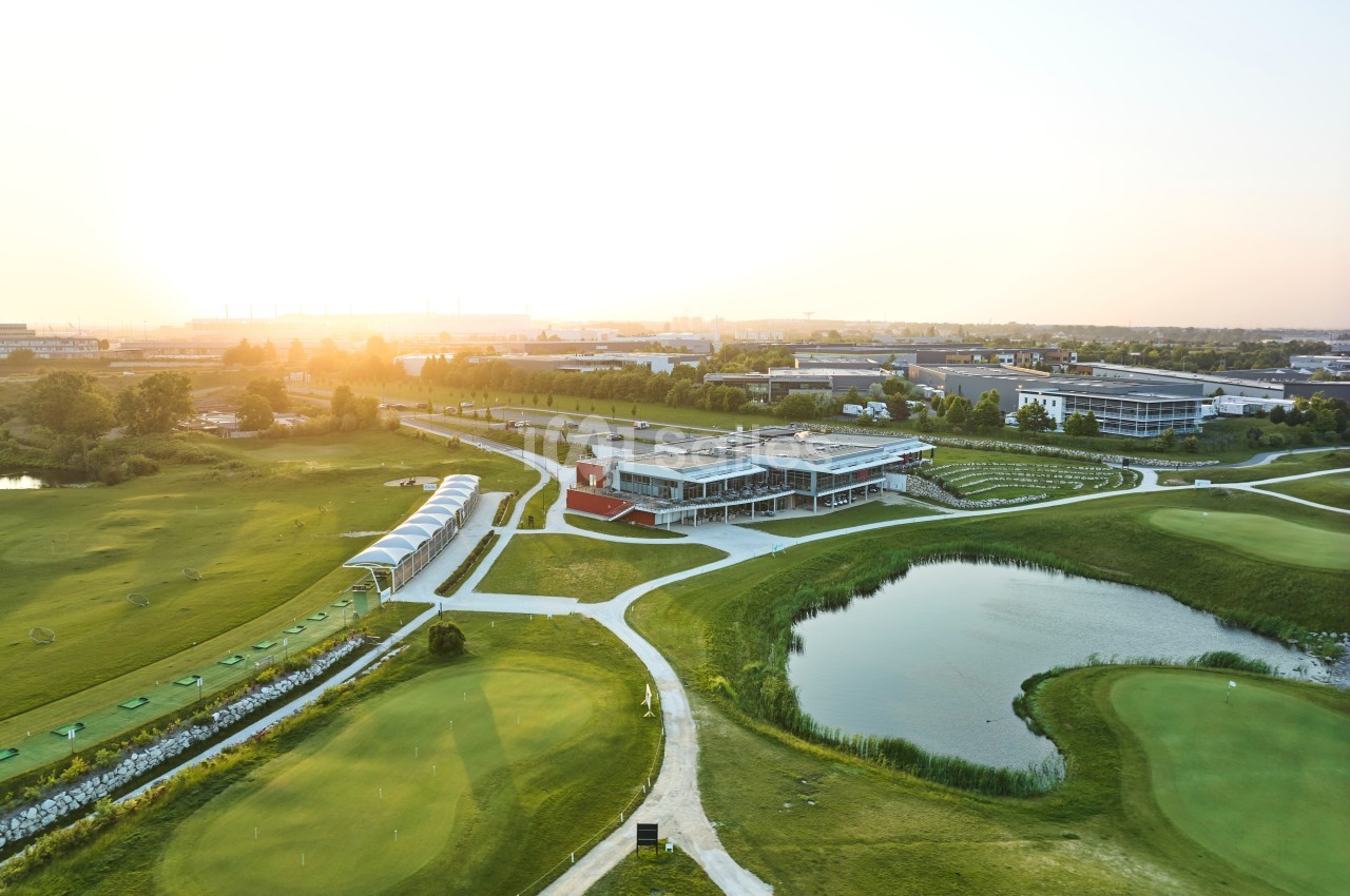 Vue aérienne d'un terrain de golf avec un bâtiment principal, des étendues d'herbe, des étangs et un coucher de soleil.