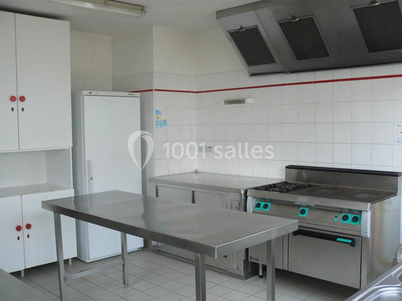 Cuisine professionnelle avec plan de travail en inox, réfrigérateur, cuisinière et hotte dans un espace carrelé blanc.