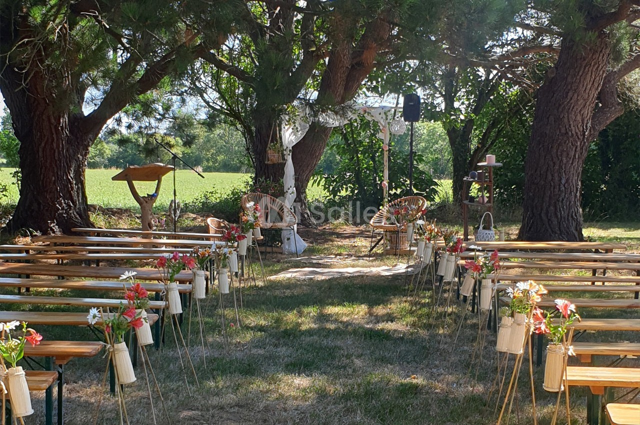 Cérémonie en plein air avec bancs décorés de fleurs, sous des arbres, face à une arche ornée de voilages blancs.