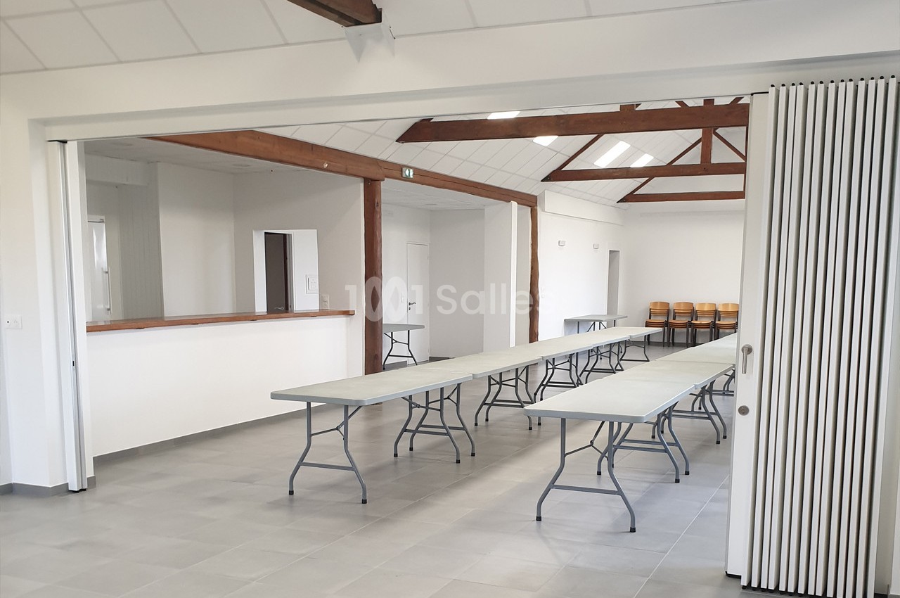 Salle lumineuse avec tables pliantes alignées, chaises empilées au fond et poutres apparentes au plafond.