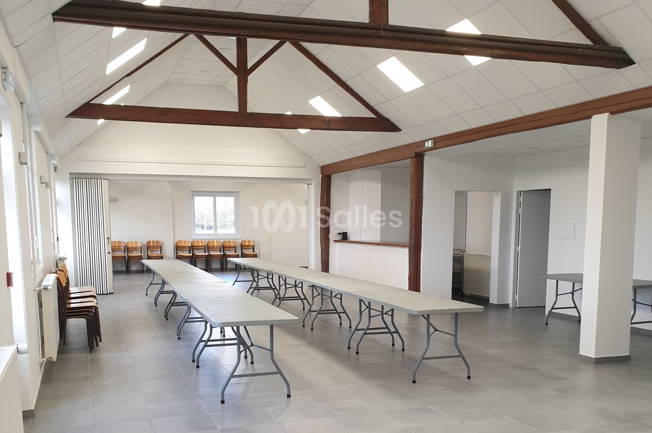 Salle lumineuse avec tables pliantes alignées, chaises contre le mur, poutres apparentes et sol en carrelage gris.