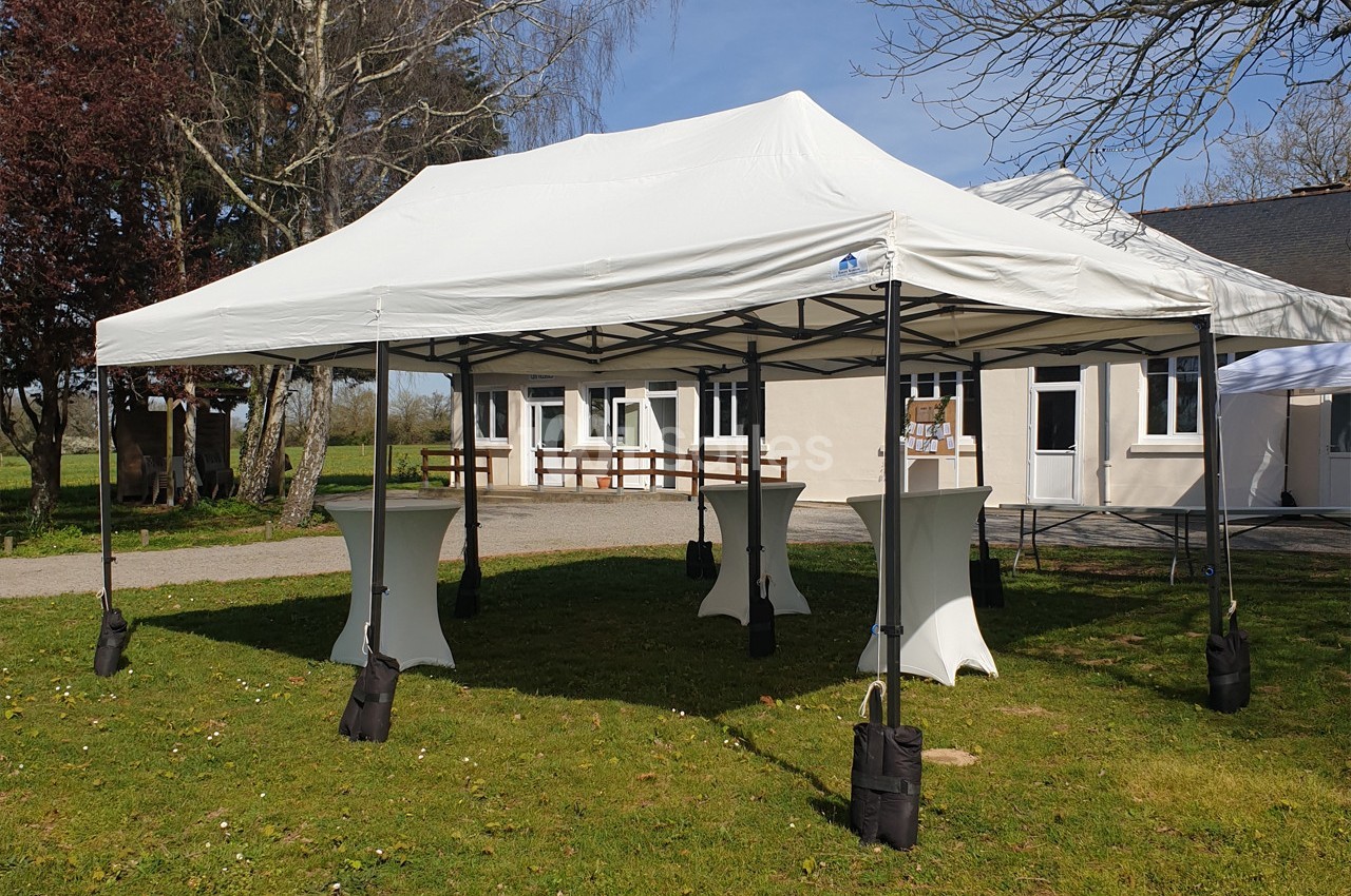 Tente blanche montée sur une pelouse, équipée de tables hautes, devant un bâtiment clair et des arbres.