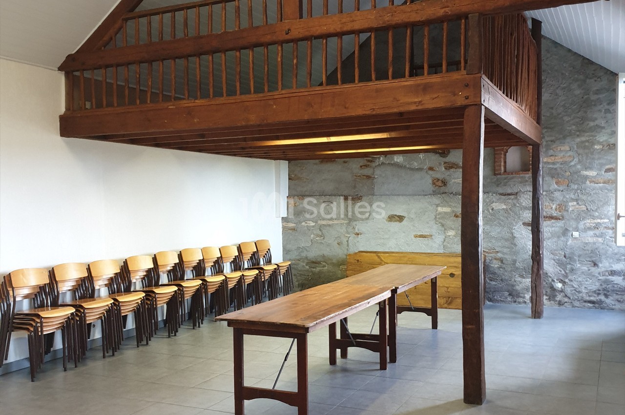 Salle avec murs en pierre et bois, mezzanine en bois, table en bois au centre et chaises empilées contre un mur.