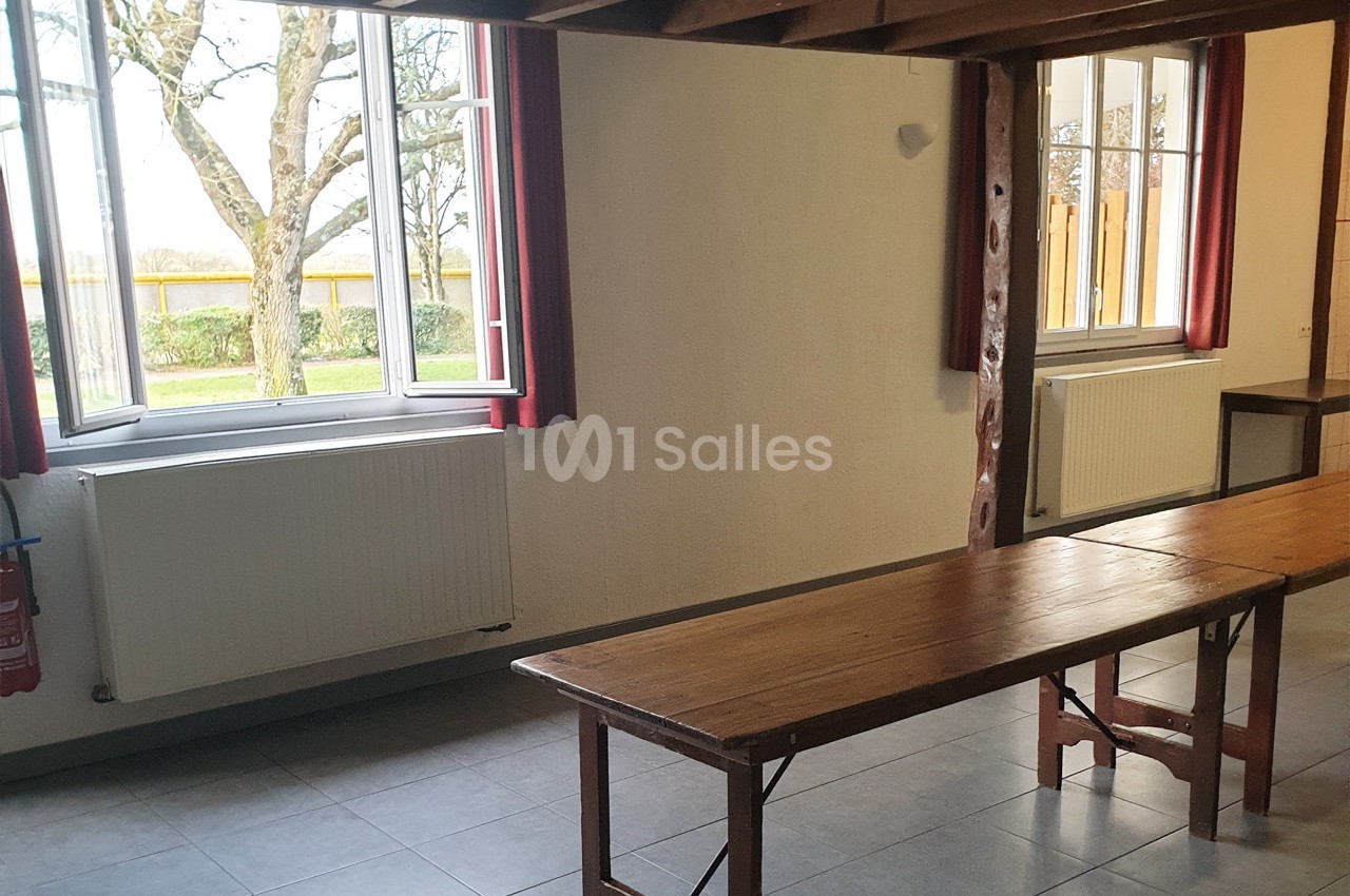 Salle lumineuse avec deux grandes fenêtres ouvertes, murs clairs, poutres en bois et tables en bois simples.