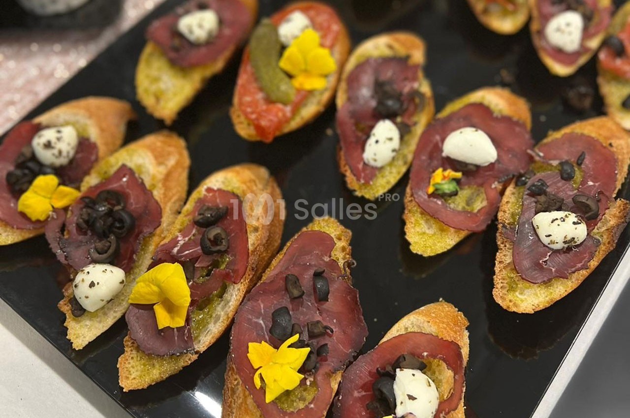 Tranches de pain garnies de viande séchée, olives noires, mozzarella, fleurs comestibles et pesto, présentées sur un plateau.