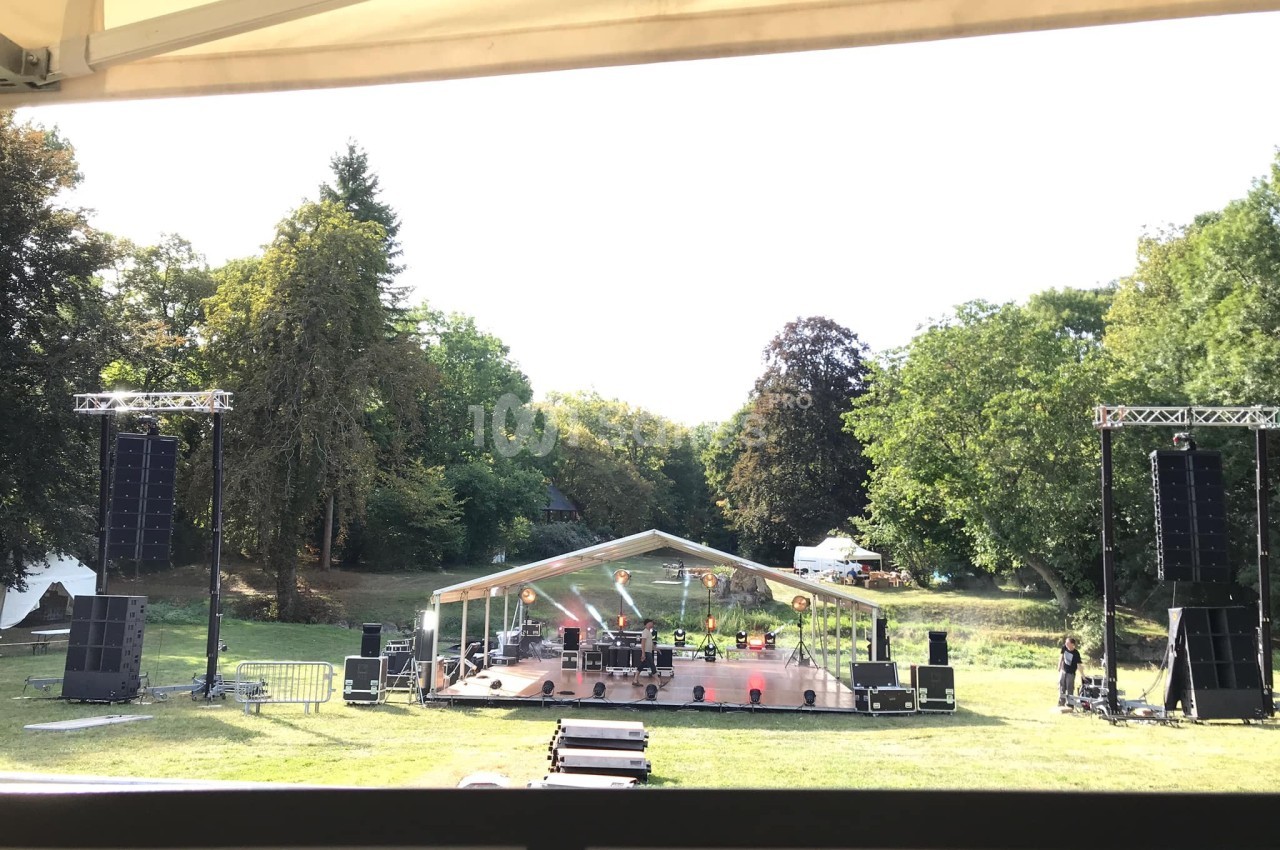 Scène extérieure avec structure couverte, équipements audio et éclairage, installée dans un parc verdoyant.