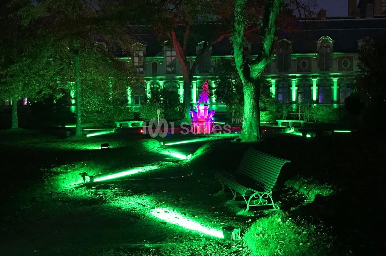 Éclairage nocturne vert illuminant un jardin avec une fontaine centrale et un bâtiment en arrière-plan.