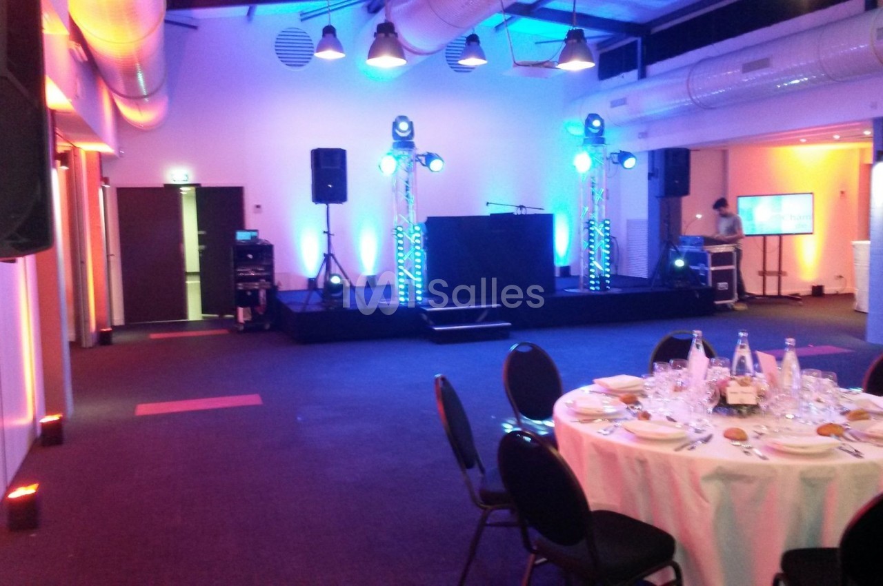 Salle aménagée pour un événement avec scène, éclairages colorés, tables dressées et équipements audio.