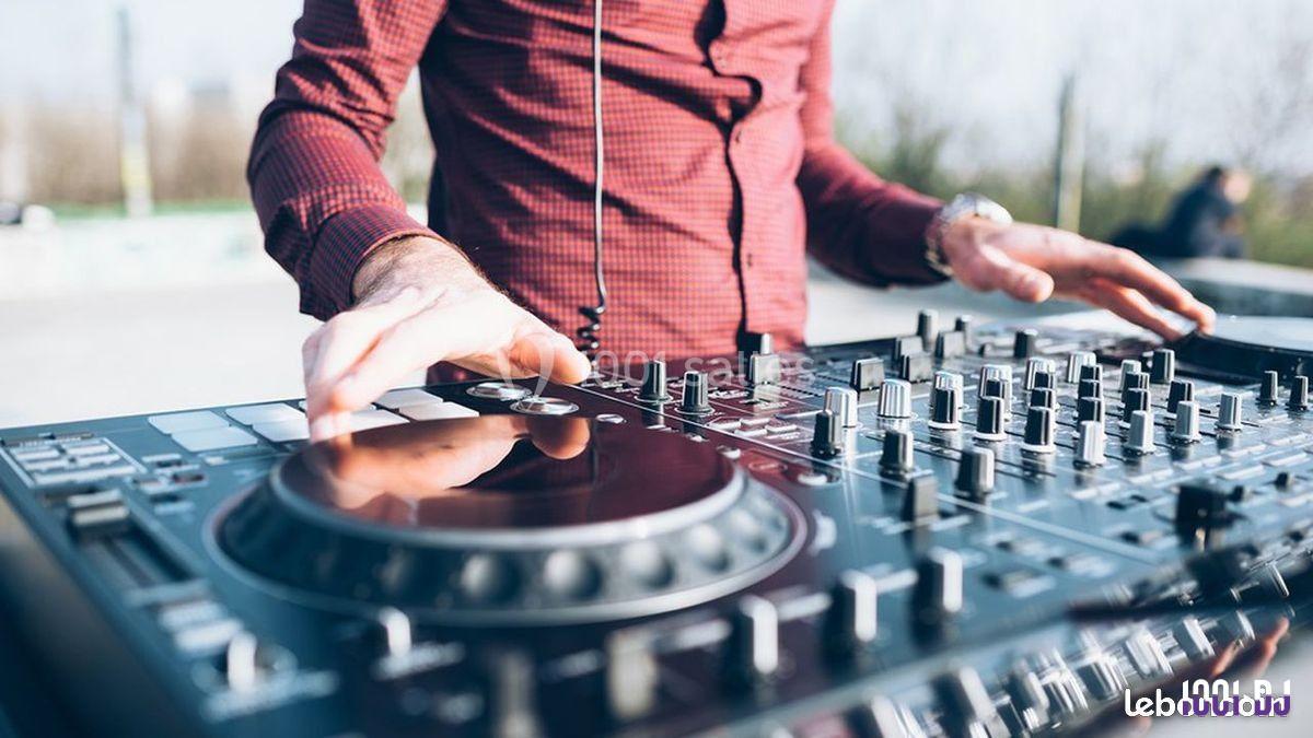Un DJ en chemise rouge mixe de la musique sur une table de mixage en extérieur.