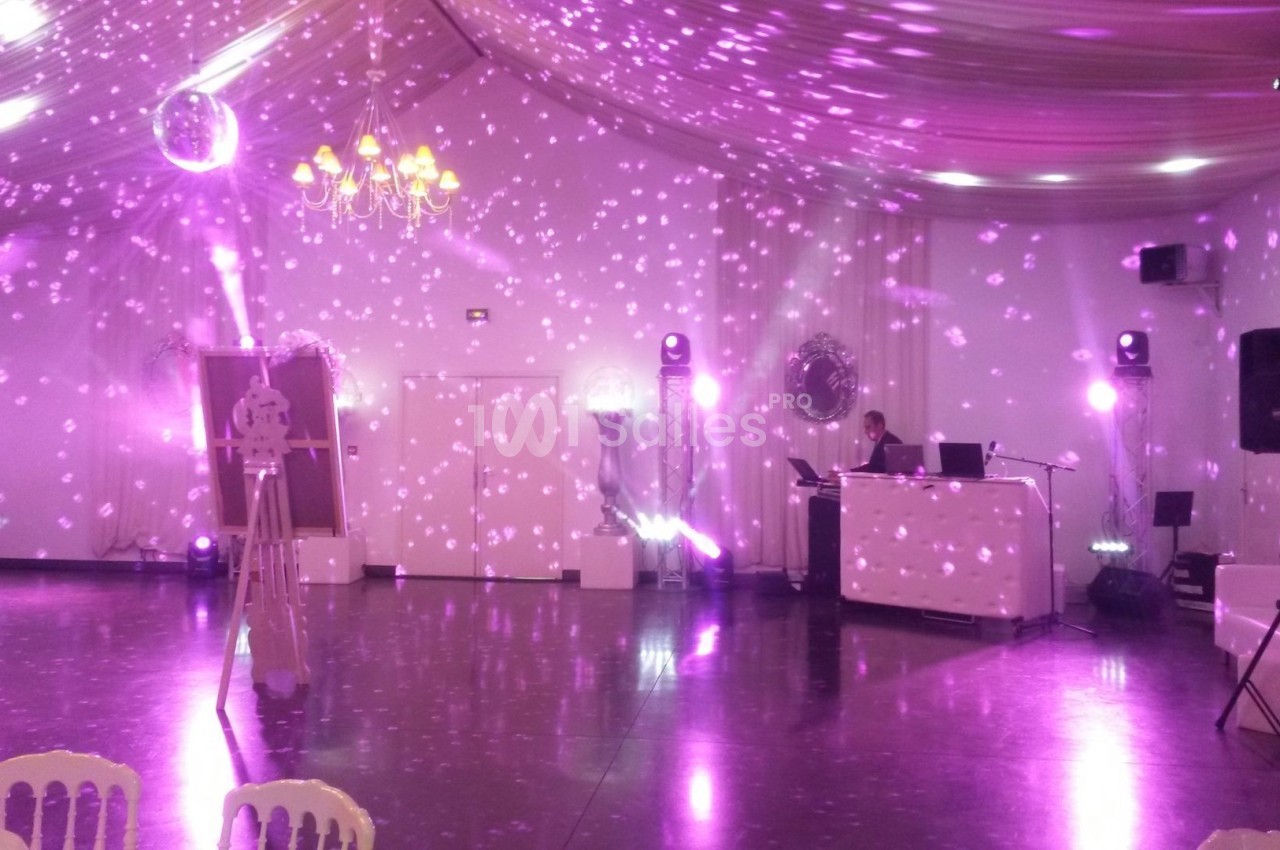 Salle de réception décorée avec des lumières violettes, un DJ à son poste et des chaises blanches au premier plan.