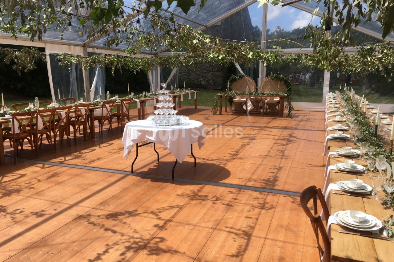 Salle de réception en plein air sous une tente transparente, décorée de tables alignées et de feuillages suspendus.