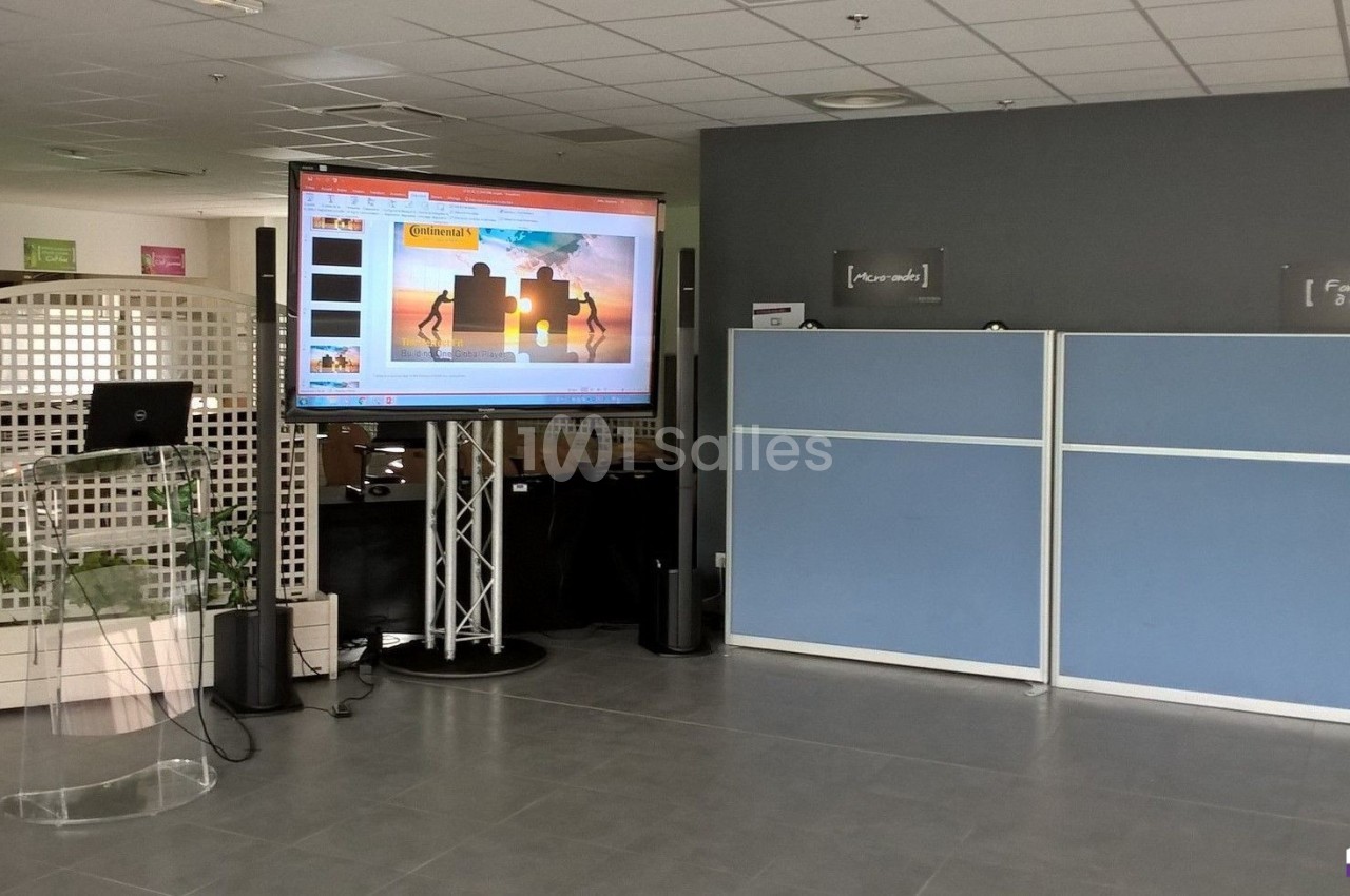 Écran affichant une présentation PowerPoint dans un espace de bureau avec cloison bleue et pupitre transparent.