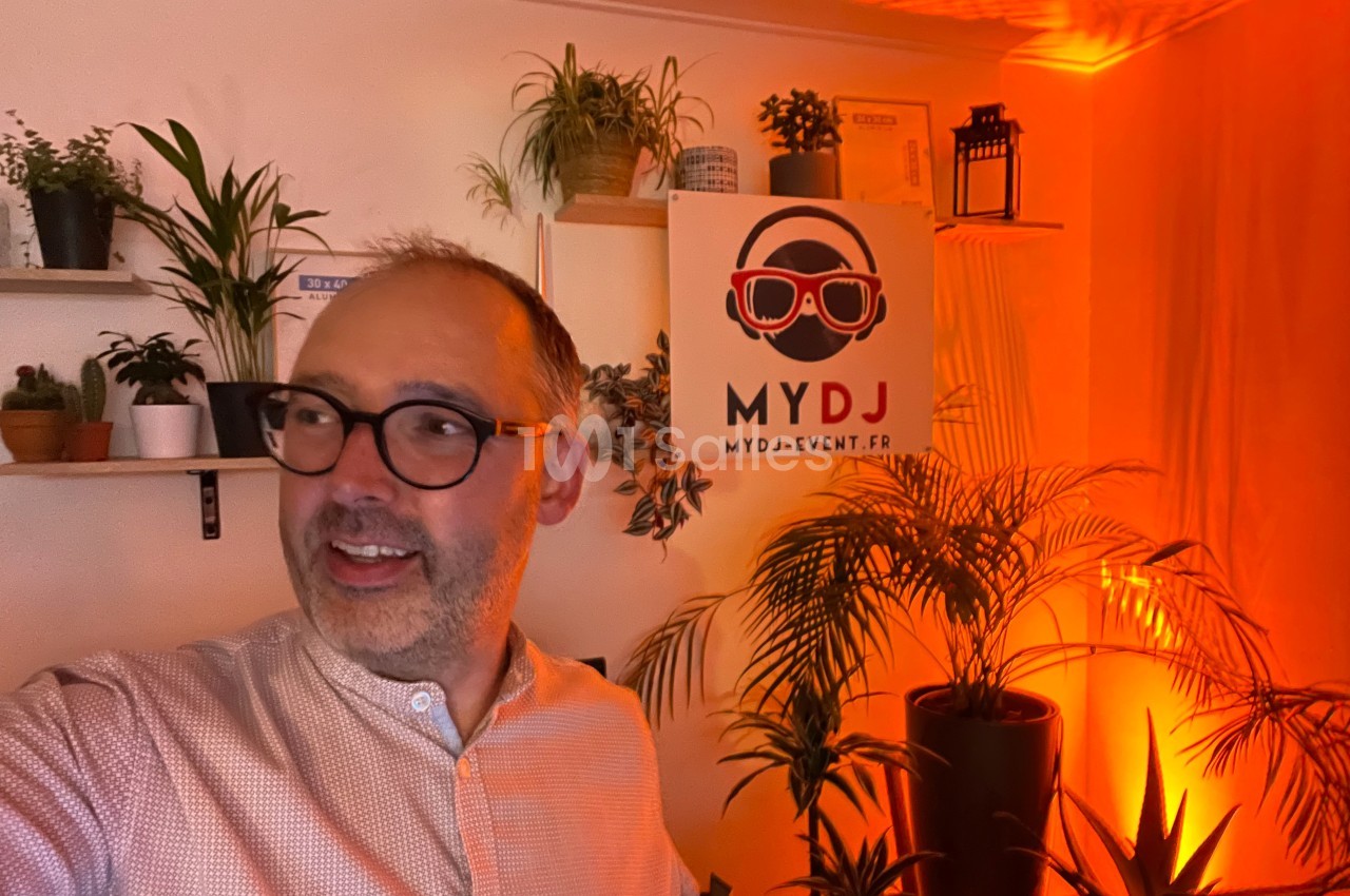 Homme souriant dans une pièce éclairée par une lumière orange, entouré de plantes et d'une affiche ’MYDJ’.