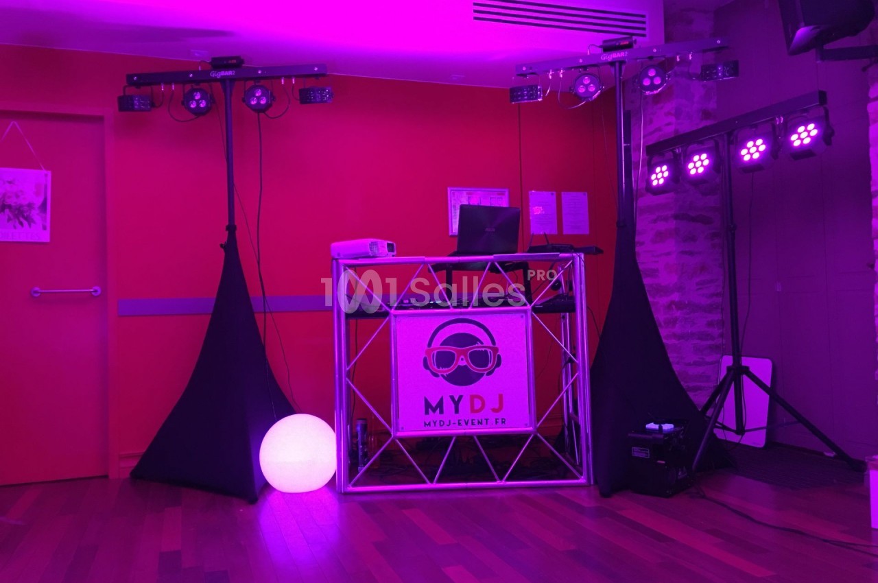 Stand de DJ avec éclairage violet, table de mixage, ordinateur portable et décorations lumineuses dans une salle.