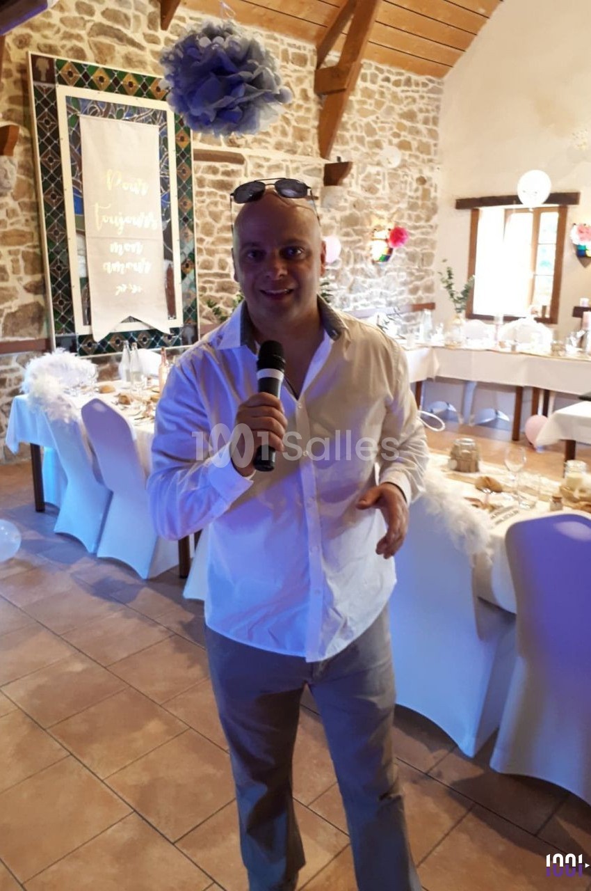 Homme debout tenant un micro dans une salle décorée pour un événement avec des tables dressées et des décorations suspendues.