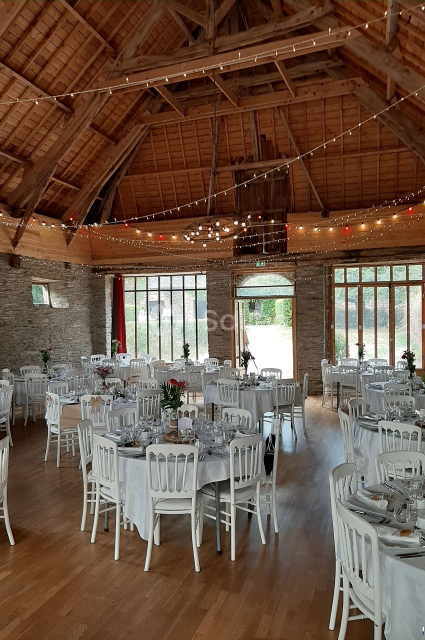 Salle de réception en pierre avec tables rondes dressées, chaises blanches et guirlandes lumineuses suspendues.