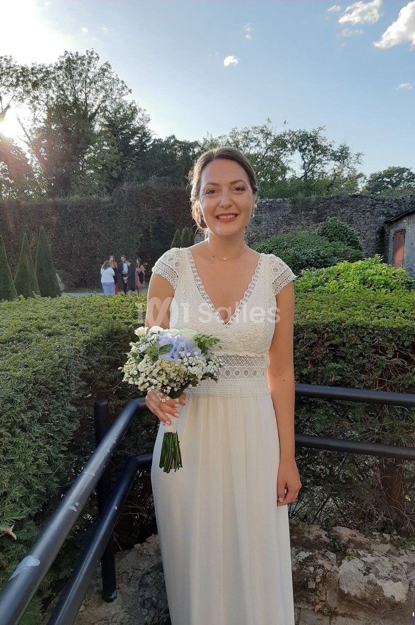 Femme en robe blanche tenant un bouquet de fleurs, debout dans un jardin avec des arbres et un mur en pierre en arrière-plan.
