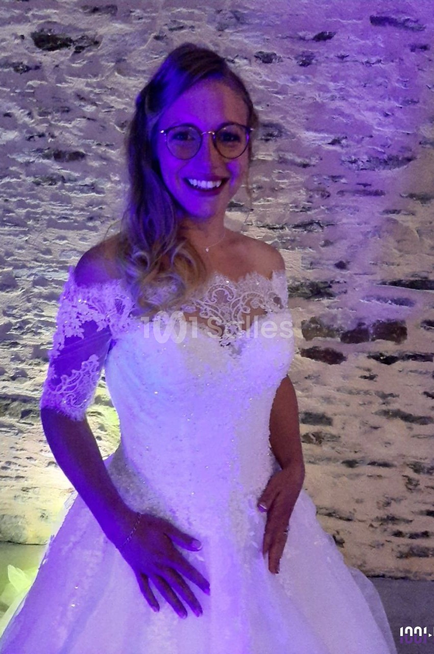 Femme souriante en robe de mariée blanche avec dentelle, posant devant un mur en pierre éclairé par une lumière violette.
