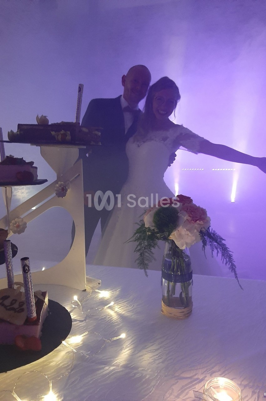 Un couple en tenue de mariage pose joyeusement devant une table décorée avec des gâteaux et un vase de fleurs.