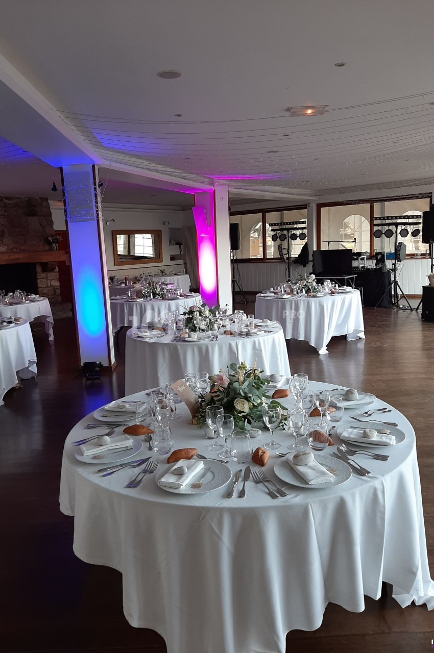 Salle de réception décorée avec des tables rondes dressées, éclairage coloré et équipement audio en arrière-plan.