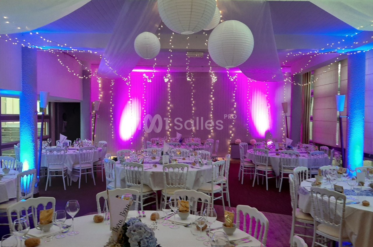Salle de réception décorée avec des guirlandes lumineuses, lanternes suspendues et tables dressées pour un événement.