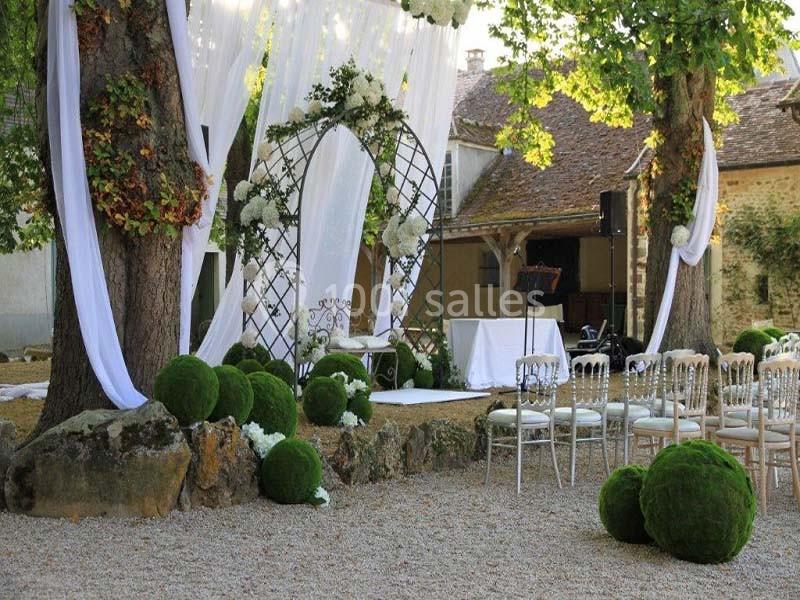 Cérémonie en plein air avec arche fleurie, chaises alignées et décorations végétales sous des arbres.