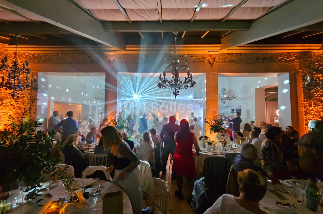 Salle de réception animée avec des invités assis et debout, éclairage coloré et jeux de lumière sur la piste de danse.