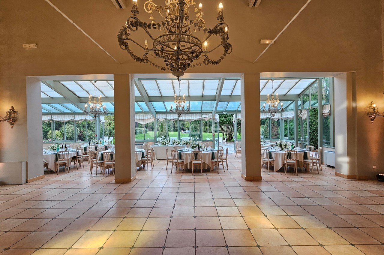 Salle de réception lumineuse avec grandes baies vitrées, vue sur un jardin, tables dressées pour un événement.