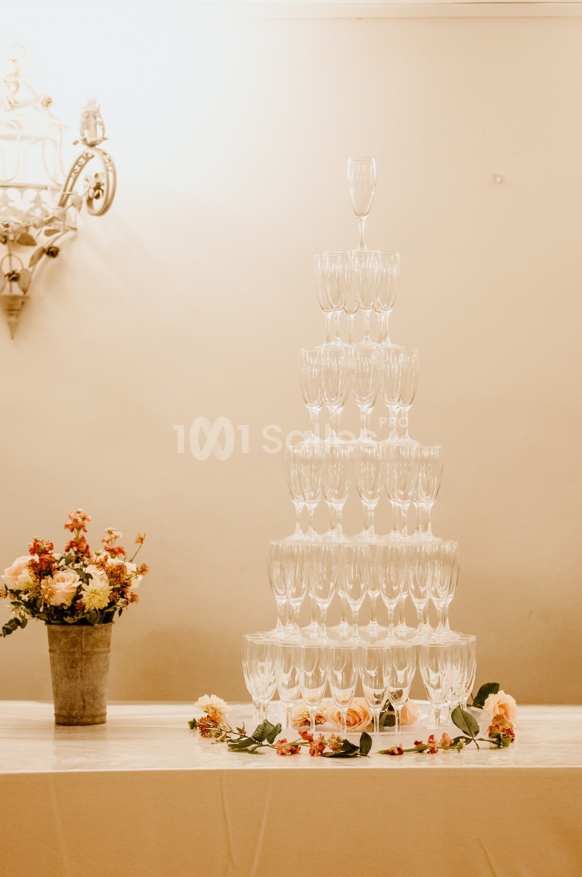 Tour de coupes en verre empilées pour un service de champagne, décorée de fleurs sur une table blanche.