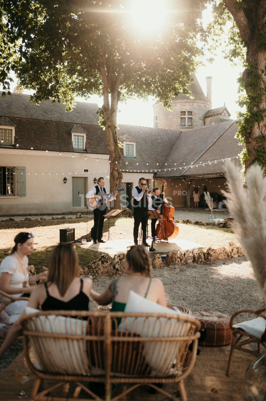 Un groupe de musiciens joue en plein air devant un bâtiment en pierre, avec des invités assis au premier plan.