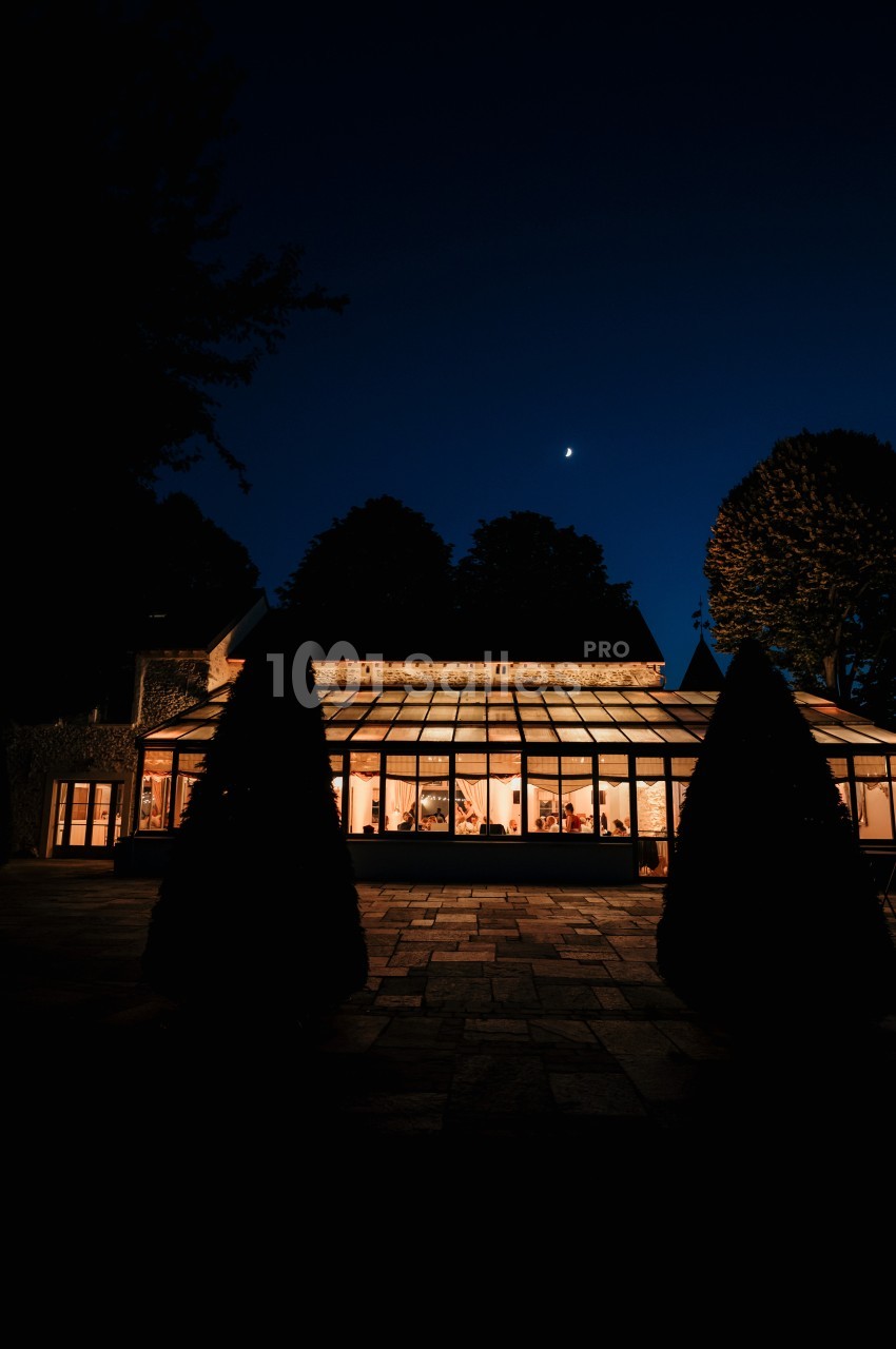 Verrière éclairée d'une maison entourée d'arbres sous un ciel nocturne avec un croissant de lune.