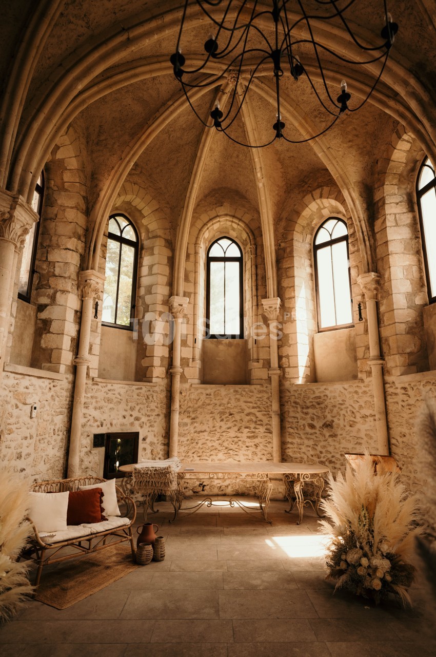 Salle voûtée en pierre avec grandes fenêtres cintrées, mobilier en fer forgé et décoration florale lumineuse.