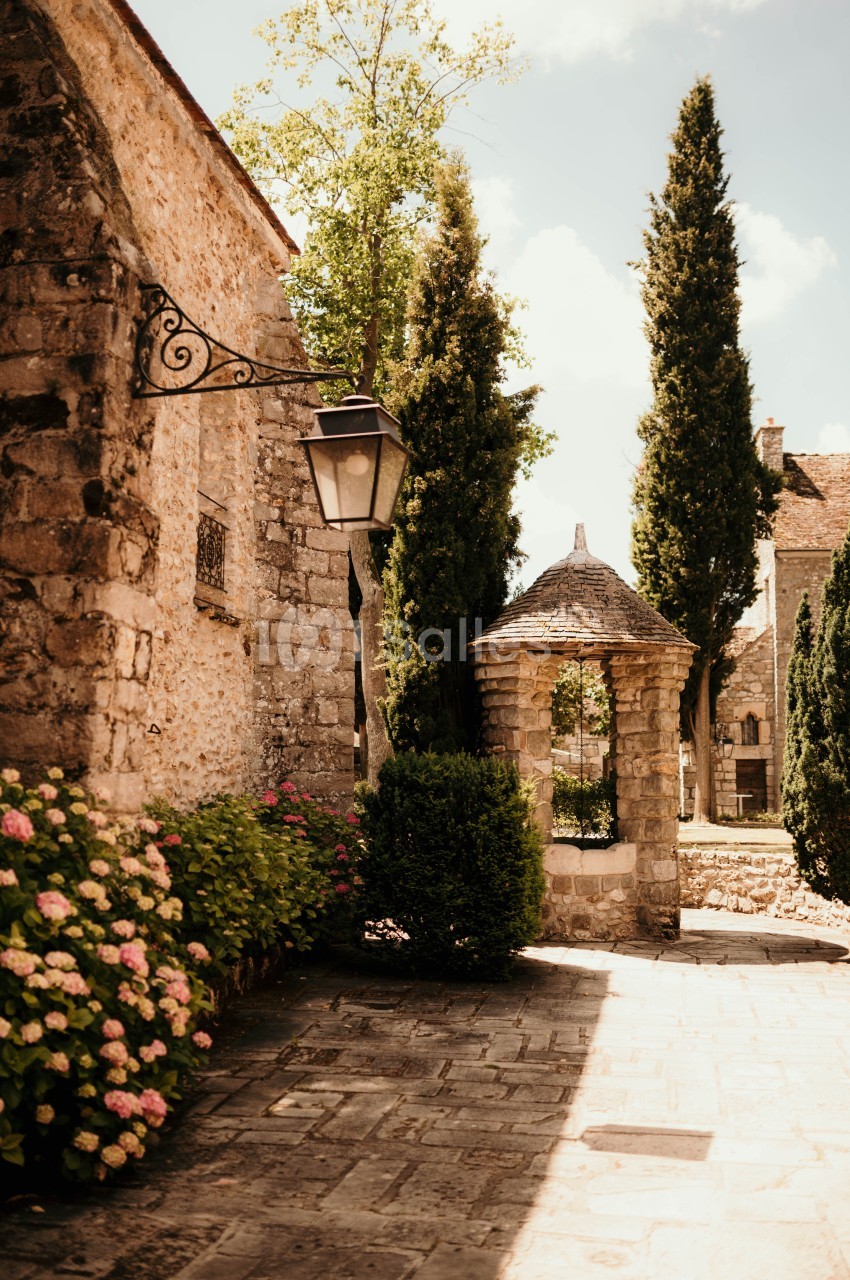 Ruelle pavée bordée de fleurs, avec un puits en pierre et des bâtiments anciens sous un ciel ensoleillé.