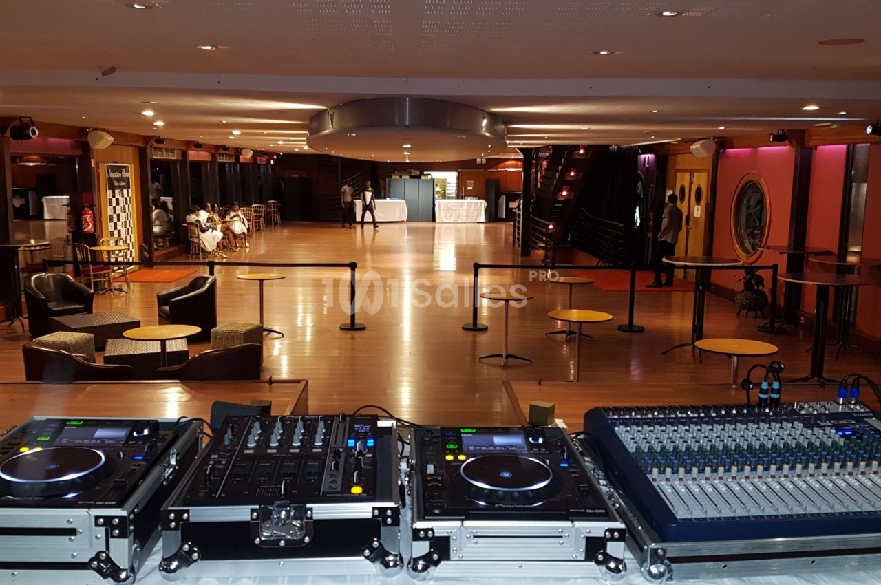 Salle spacieuse avec tables et chaises, vue depuis une régie équipée de platines DJ et d'une console de mixage.